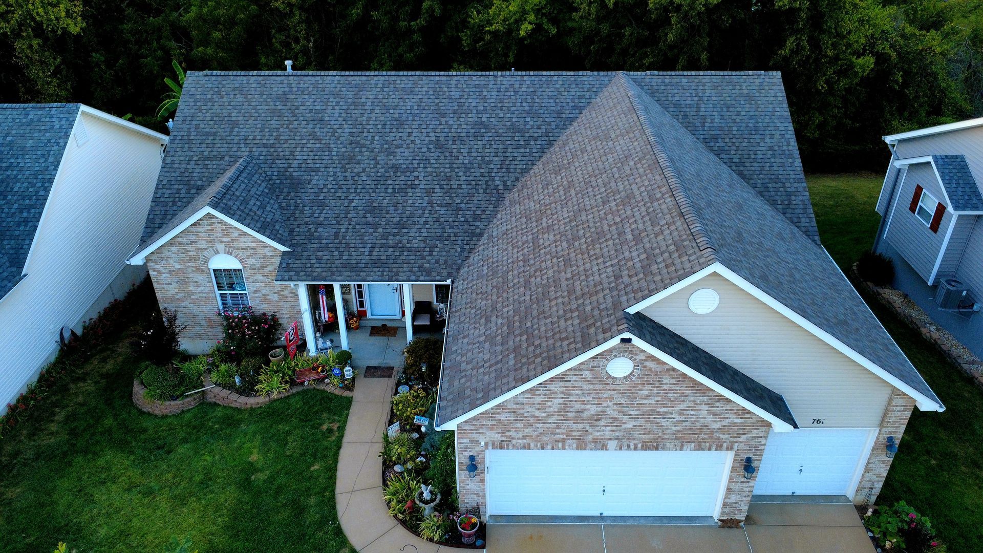 An aerial view of a suburban brick house with a dark gray shingled roof, a two-car garage, and a landscaped front yard.