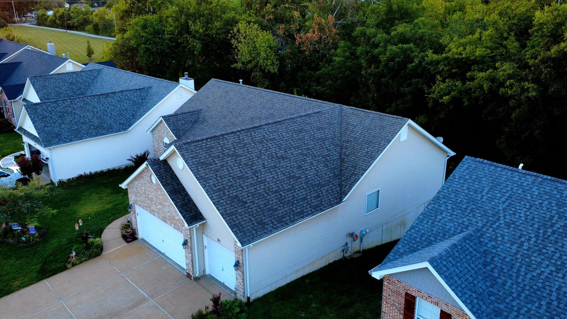 Aerial view of suburban houses with grey roofs and driveways bordering a line of dense green trees.