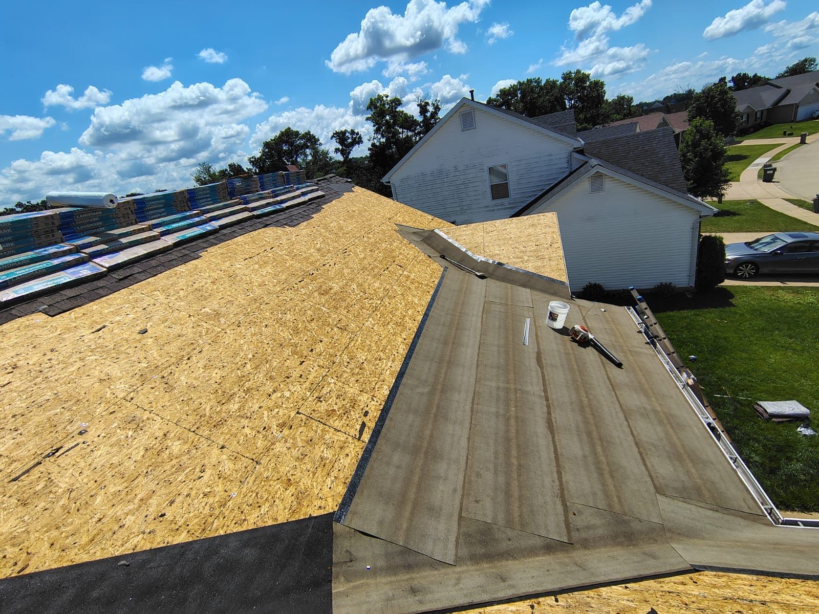 A partially re-roofed house with sections of exposed plywood decking and gray underlayment under a sunny blue sky.