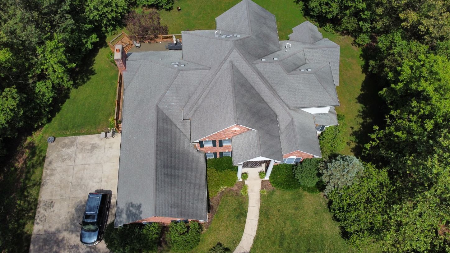 An aerial view of a large residential house with a gray shingled roof, a paved driveway, and lush surrounding trees.