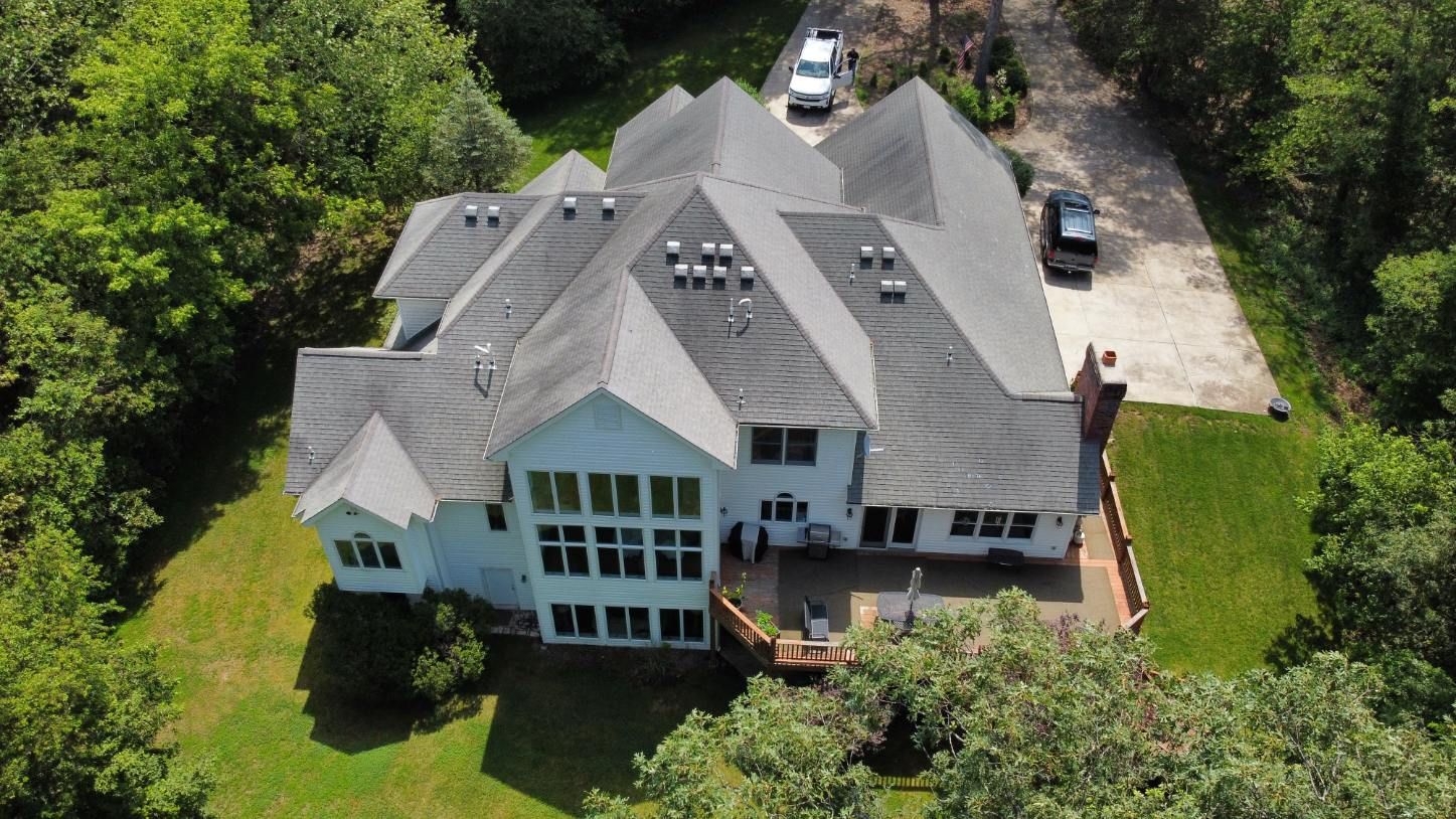 An aerial view of a large white two-story house with a grey roof, surrounded by trees, a driveway, and a backyard patio.