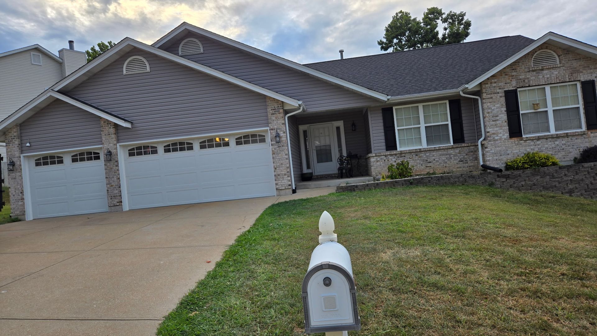 A one-story suburban home with a gray vinyl and brick exterior, three-car garage, and a mailbox in the front yard.