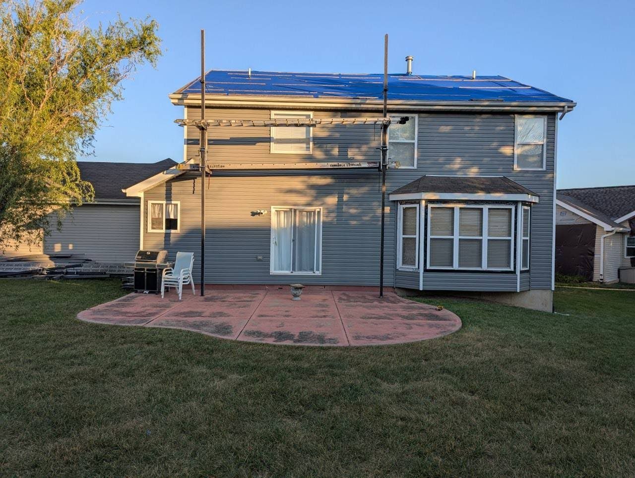 Back of a gray two-story suburban house with a blue tarp on the roof, scaffolding, and a patio with a chair.