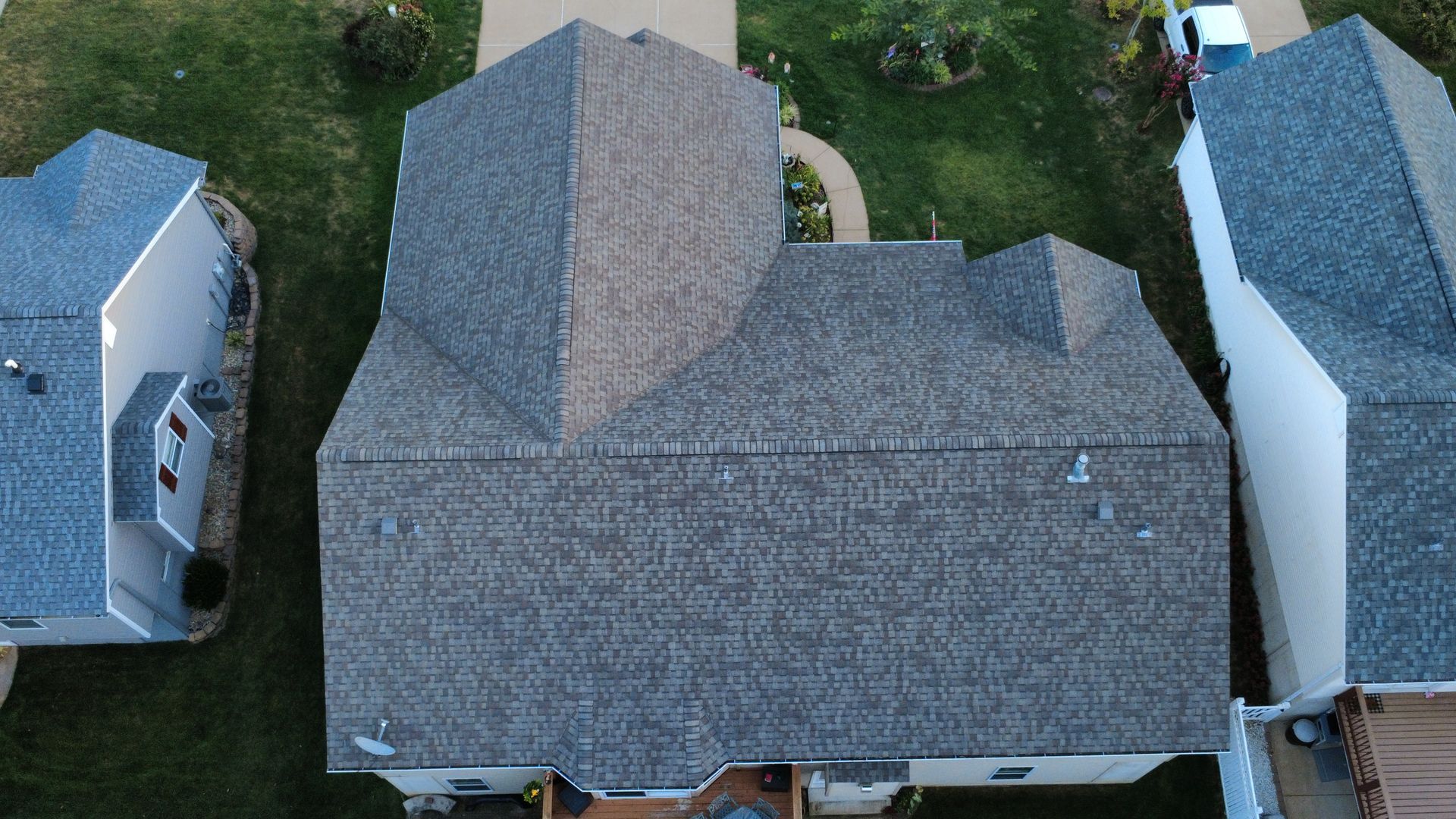 An aerial view of a gray-shingled residential house roof surrounded by green lawns and neighboring homes.