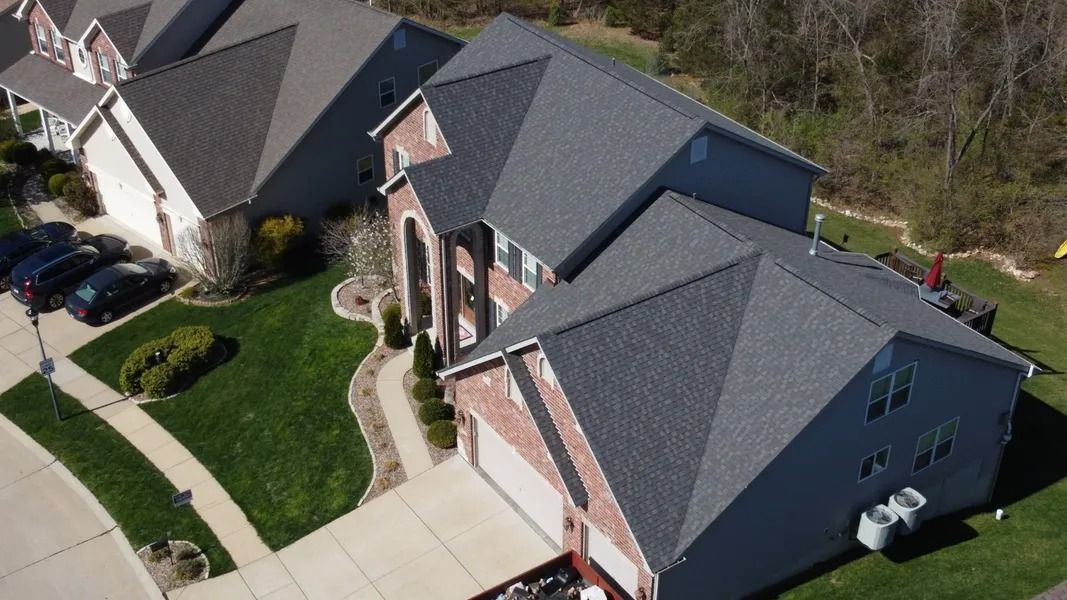 An elevated, angled view of a suburban brick house with a dark grey roof and a manicured lawn on a sunny day.