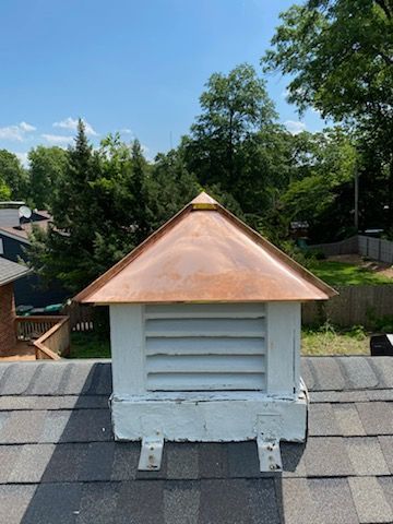 A white roof cupola with a polished copper pyramid roof, situated on a dark shingled roof with trees in the background.