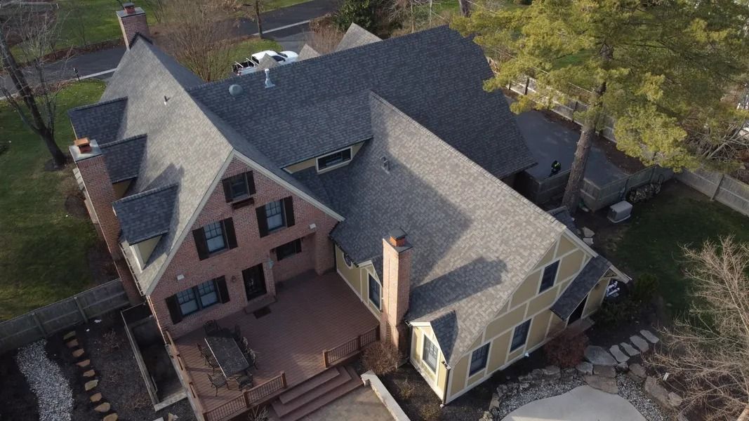 Aerial view of a large residential brick and siding house with a complex shingled roof, patio, and surrounding yard.