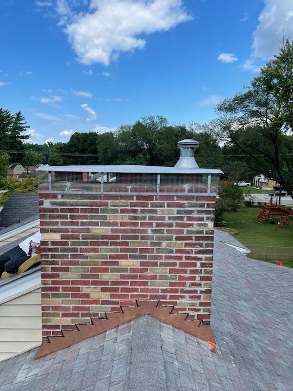 Brick chimney on a shingled roof with blue sky and trees in the background