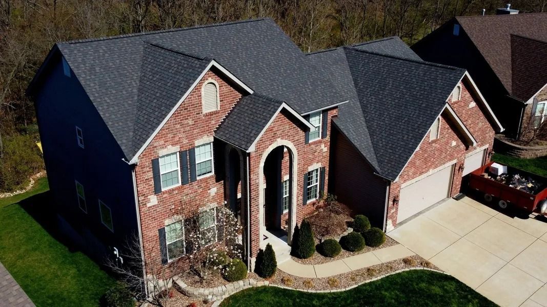 An aerial view of a two-story brick suburban house with a dark shingled roof, arched entryway, and attached garage.