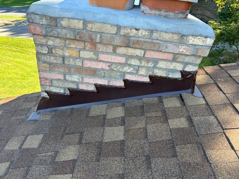 Damaged brick chimney base with missing mortar and lifted shingles on a rooftop