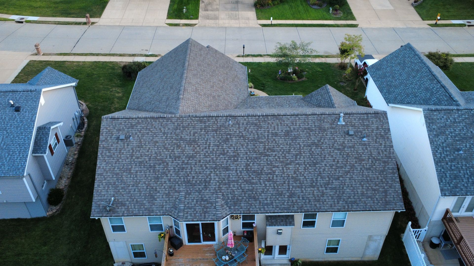 A high-angle aerial view of a gray shingled residential house roof, surrounded by green lawns and a paved suburban street.