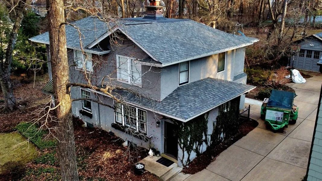 An aerial view of a two-story gray house with a gray shingled roof, surrounded by trees and a concrete driveway.