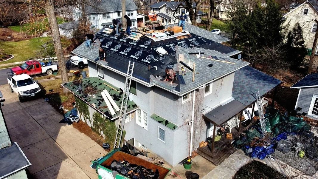 Aerial view of a residential roof replacement with workers, construction materials, and a dumpster in the driveway.