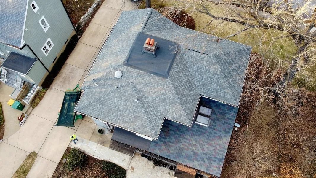 An aerial view of a gray-shingled residential roof with a central chimney and a dumpster parked in the driveway below.
