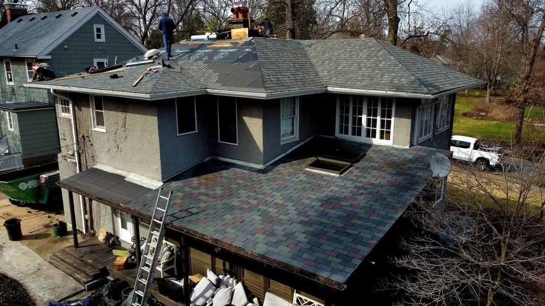 An aerial view of workers repairing the shingled roof of a gray multi-story house on a sunny day.