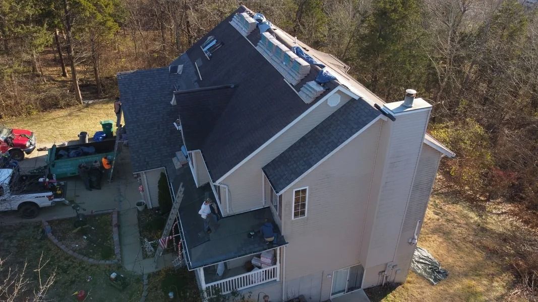 Aerial view of a tan two-story house undergoing roof repairs, with two work trucks parked in the driveway below.