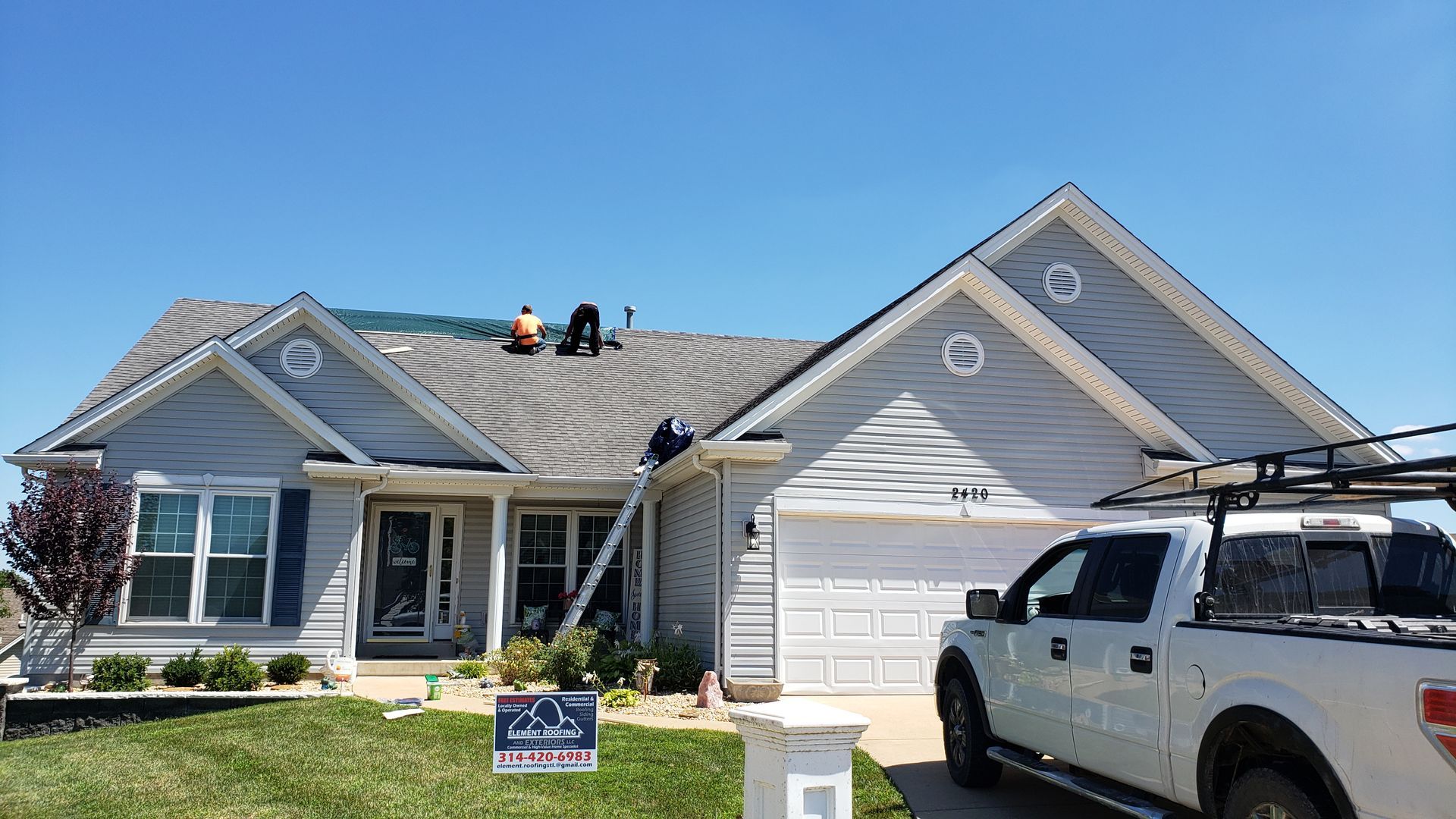 Suburban house with workers on the roof, a white truck in the driveway, and a lawn sign in front