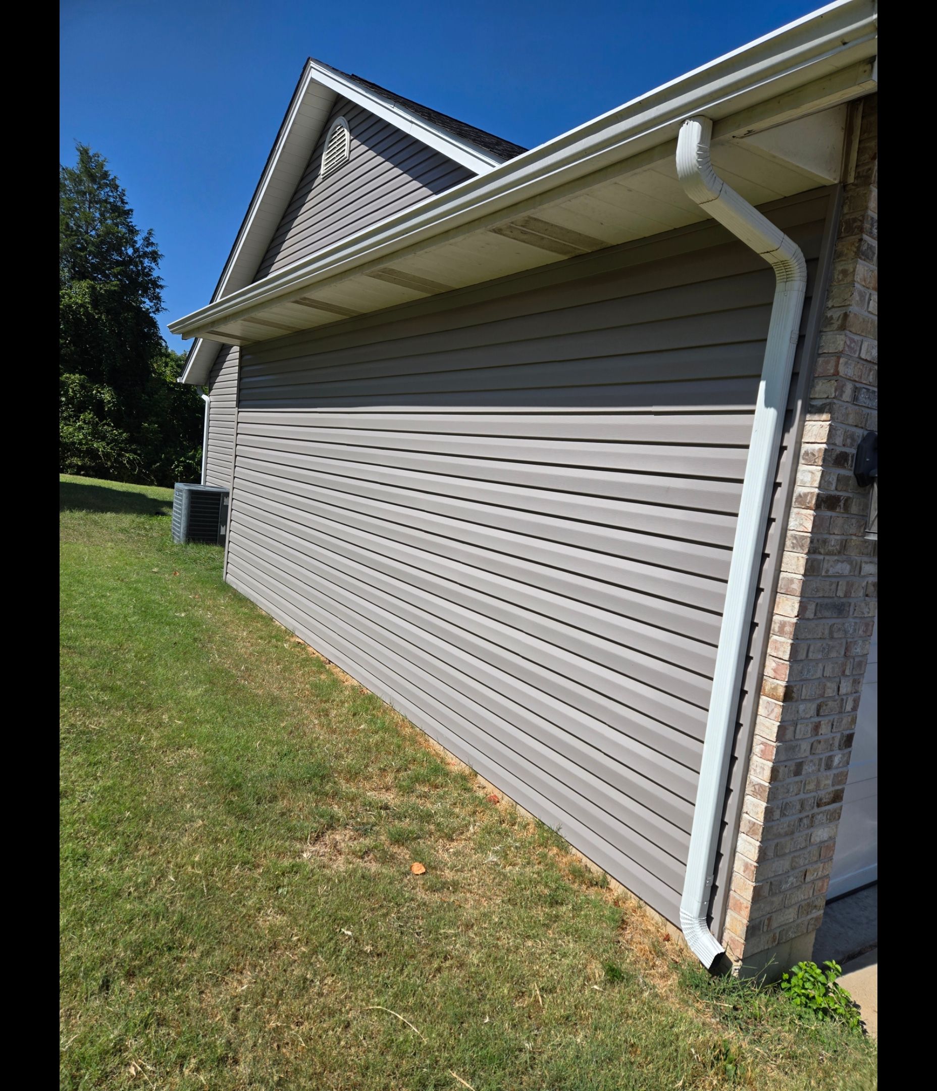Side view of a house exterior featuring grey horizontal vinyl siding, a white gutter downspout, and a brick corner.