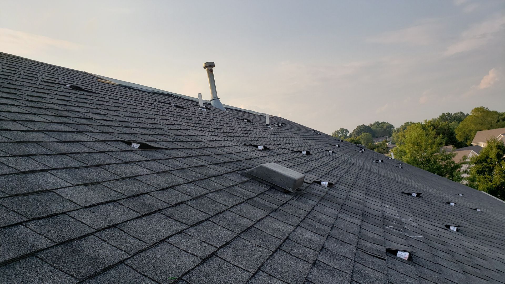 Dark shingled roof with skylights and a vent under a hazy sky, trees and houses in the distance