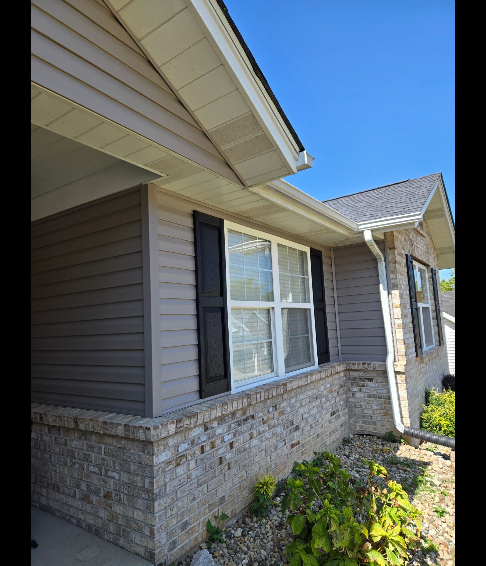 An exterior view of a house with tan siding, stone masonry on the lower half, and black window shutters.