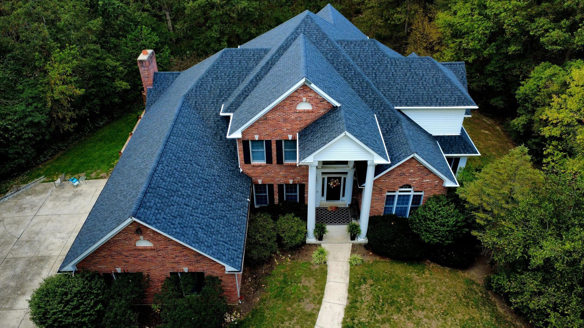 An aerial view of a two-story red brick house with a blue shingled roof, surrounded by trees and a concrete driveway.