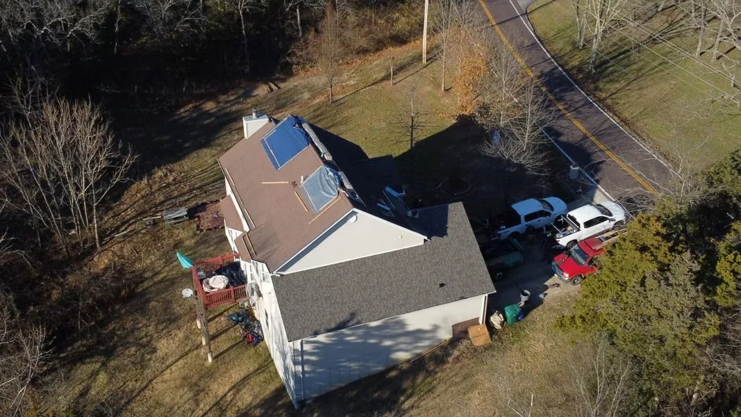 Aerial view of a house with a blue roof, parked vehicles, and trees in a rural yard.
