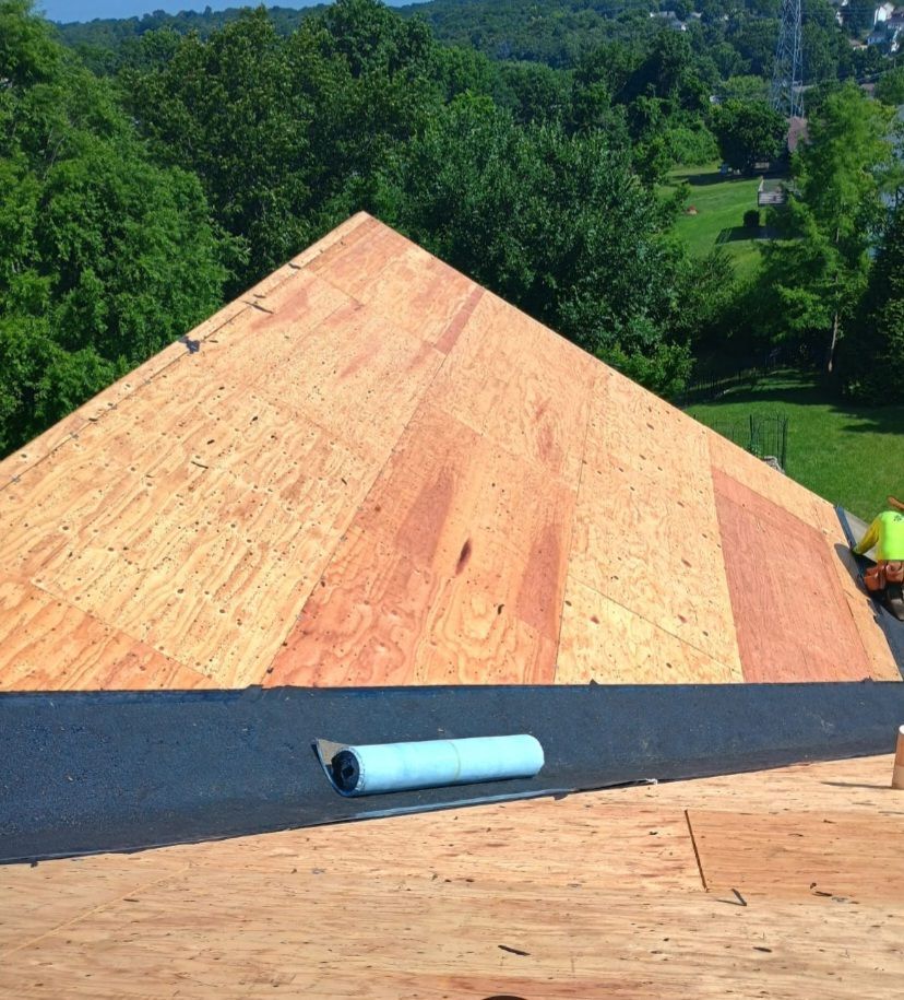 A worker installs black roofing underlayment over plywood roof decking on a sunny day.