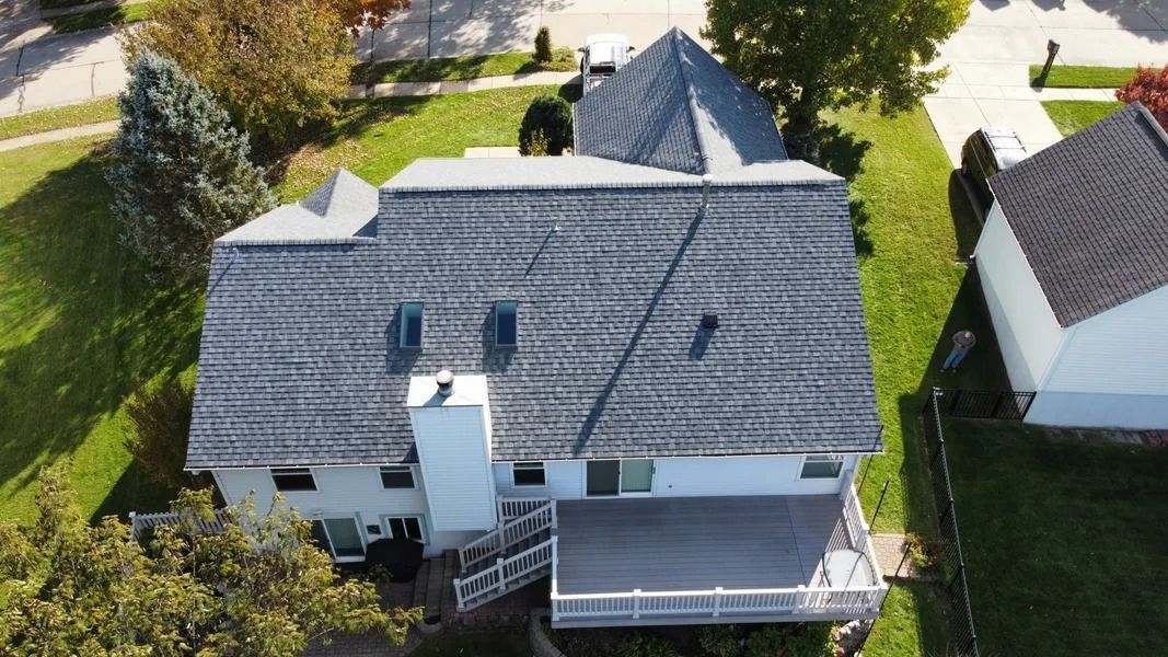 An aerial view of a white two-story house with a grey shingled roof, a chimney, and a wooden back deck on a grassy lot.