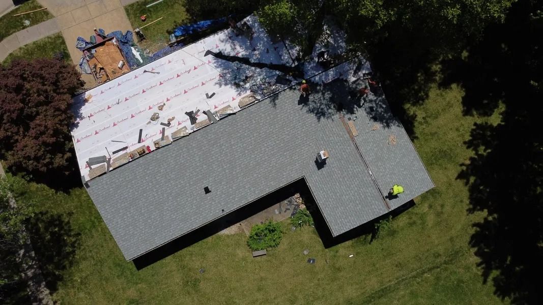 An aerial view of a residential roof under repair, with one side covered in white underlayment and the other in gray shingles.