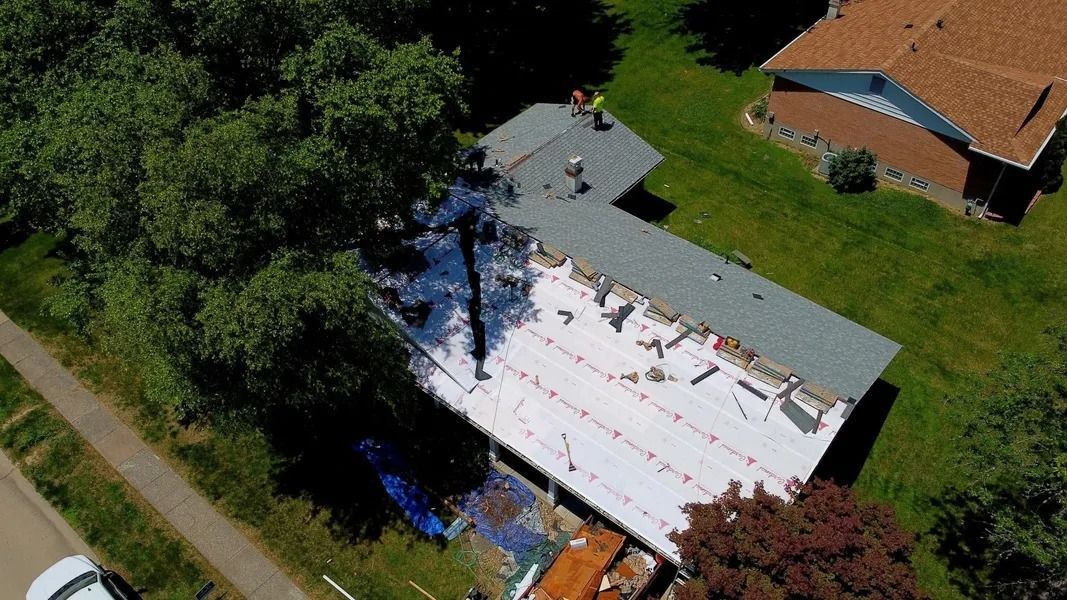 Aerial view of workers replacing a residential roof, with half the roof covered in white underlayment and half in shingles.
