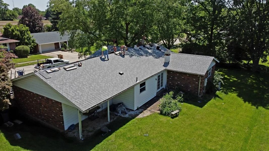 Aerial view of workers installing new grey shingles on the roof of a brick and white-sided suburban house.