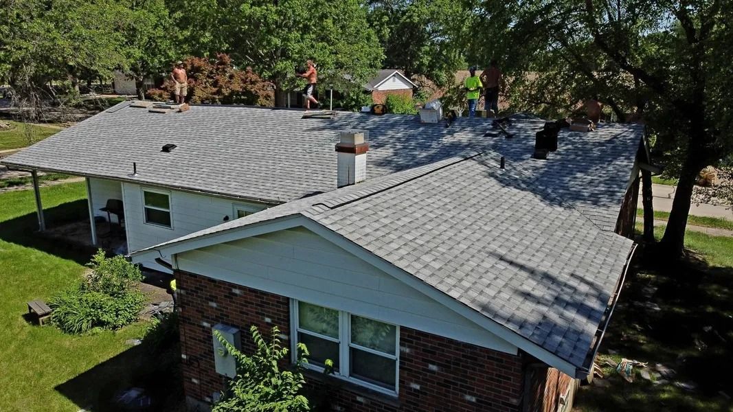 Workers stand on the roof of a house with light-colored shingles and a brick exterior during a sunny day.