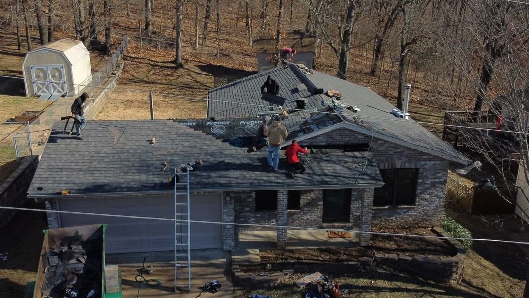 Roofers working to replace gray shingles on a residential home with a detached shed and a dumpster nearby.
