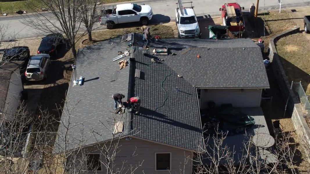 Aerial view of workers replacing gray asphalt shingles on a residential house roof with parked vehicles nearby.