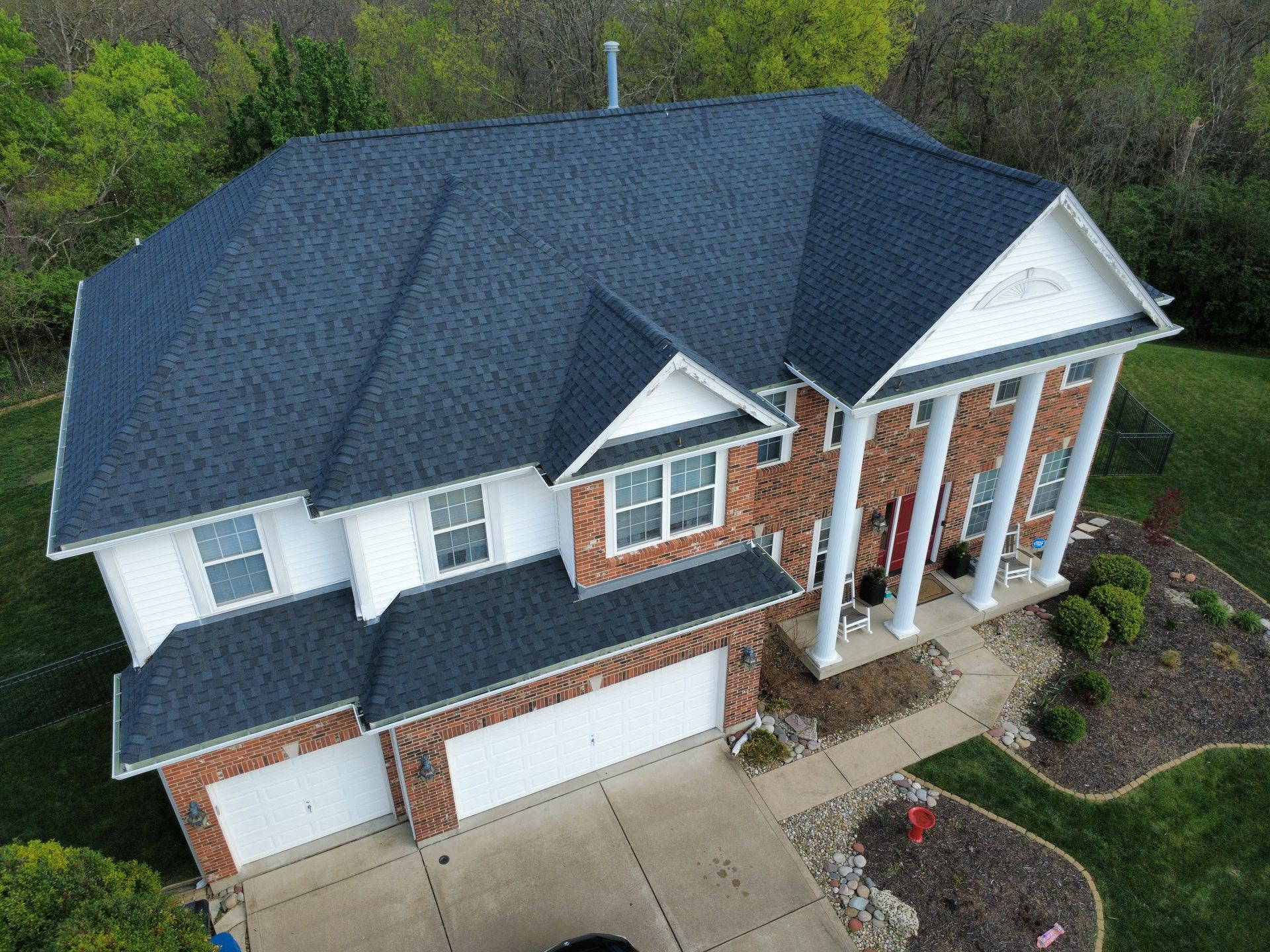 Aerial view of a two-story brick house with a dark blue roof, white columns, and a three-car driveway.