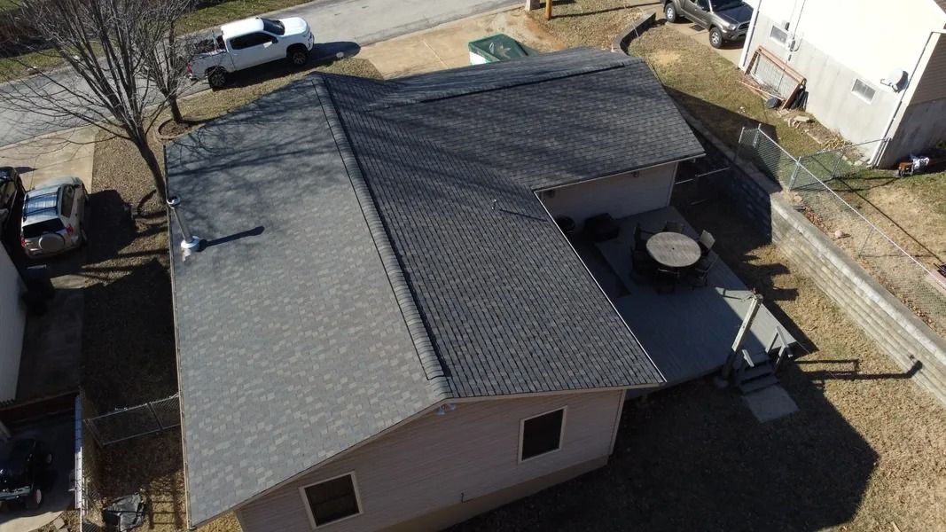 An aerial view of a tan, L-shaped residential house with a dark gray shingled roof, surrounded by a yard and driveway.