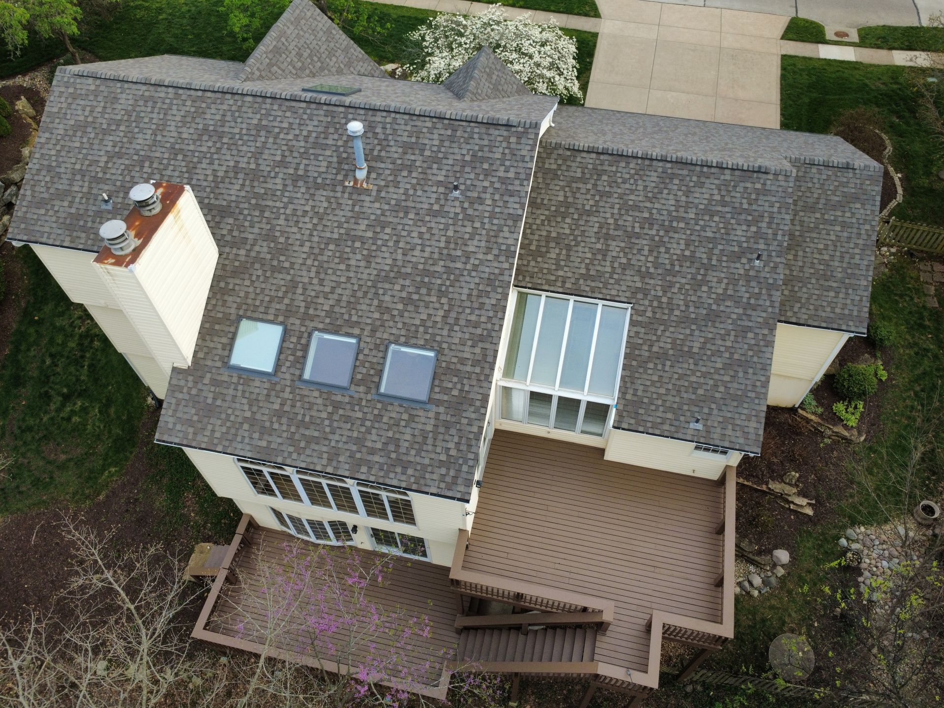 Aerial view of a gray-roofed house with skylights, a chimney, and a wooden deck surrounded by trees.