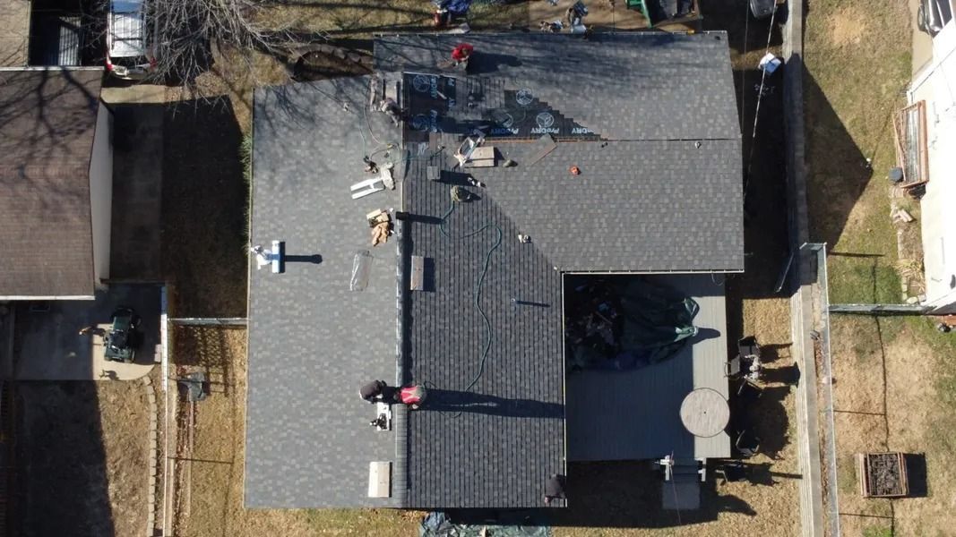 An aerial view shows workers on a gray asphalt shingle roof, with a yard and surrounding structures visible.