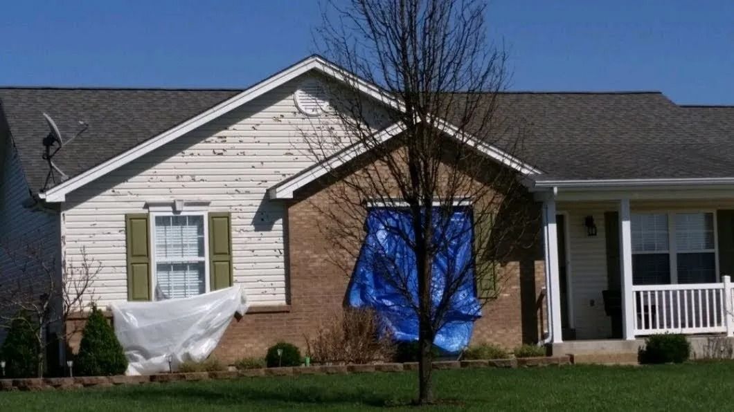 A brick house with storm-damaged siding and blue and white tarps covering two front windows under a bright blue sky.