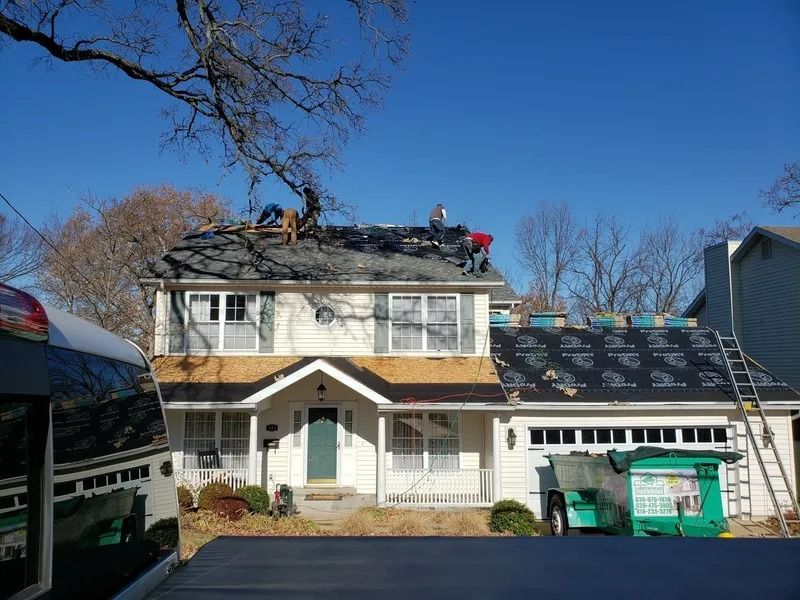 House roof being repaired with workers on top, beside a green dumpster under a clear blue sky