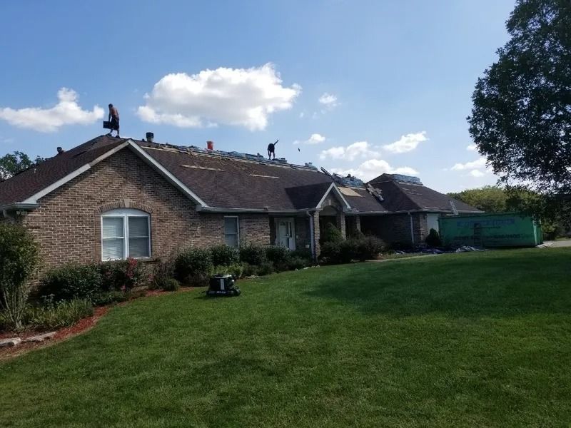 Workers on a roof during a home renovation project with a green dumpster parked in the yard.