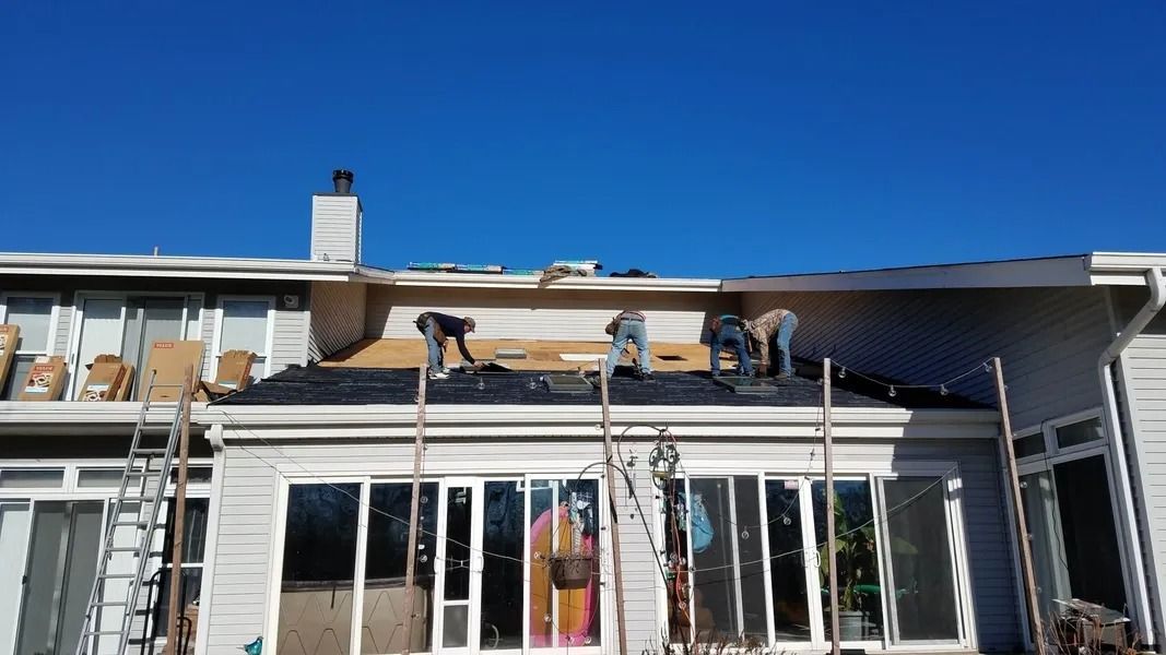 Four workers replace shingles on a roof section above a patio with large sliding glass doors under a clear blue sky.