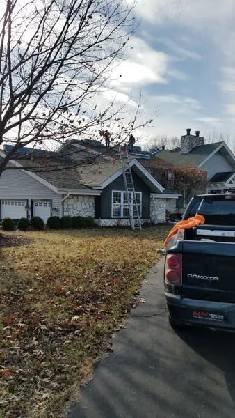 Construction workers on the roof of a suburban house with a ladder leaning against it and a pickup truck in the driveway.