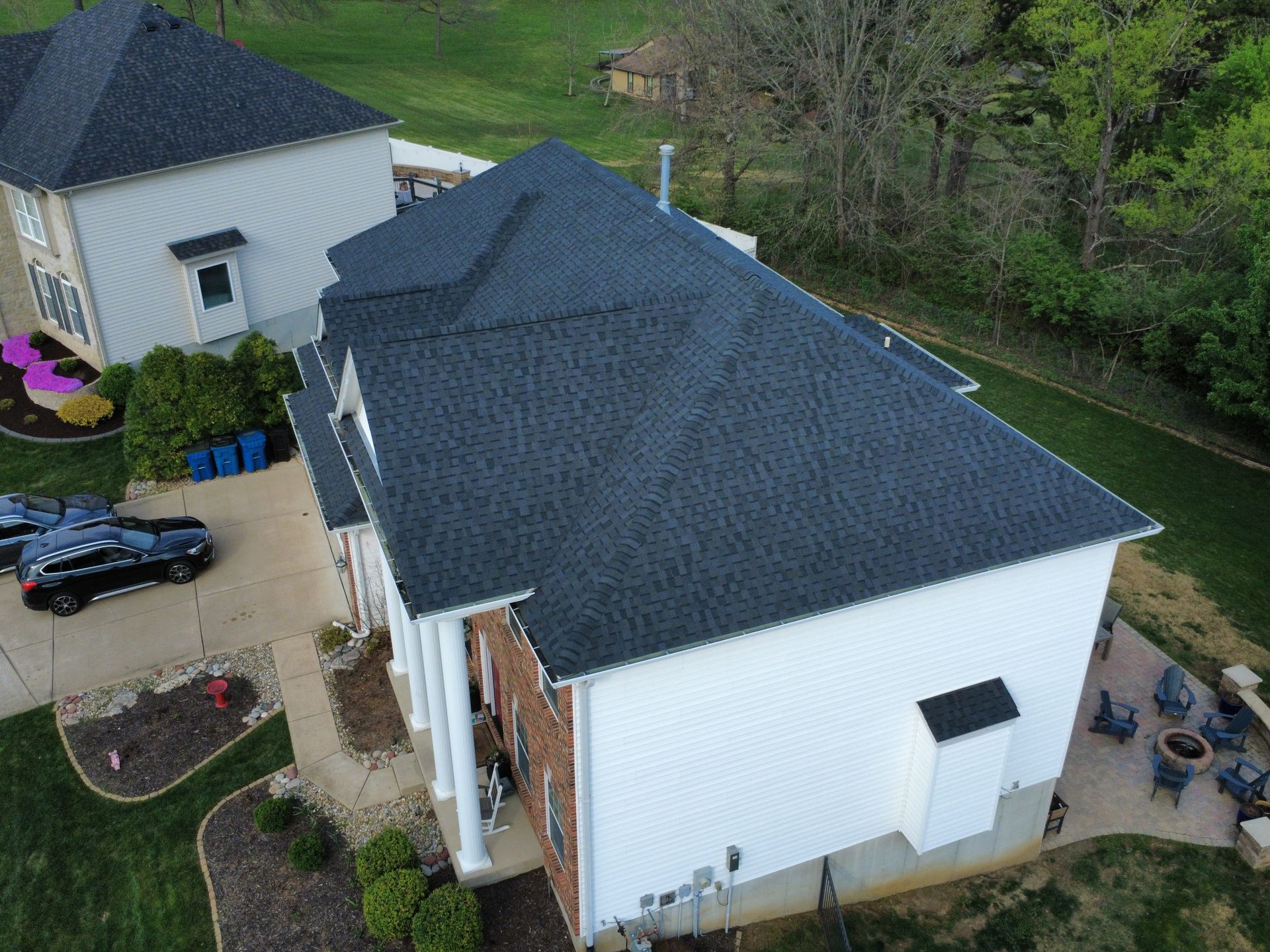 Aerial view of a white house with a dark roof and small yard beside a wooded area.