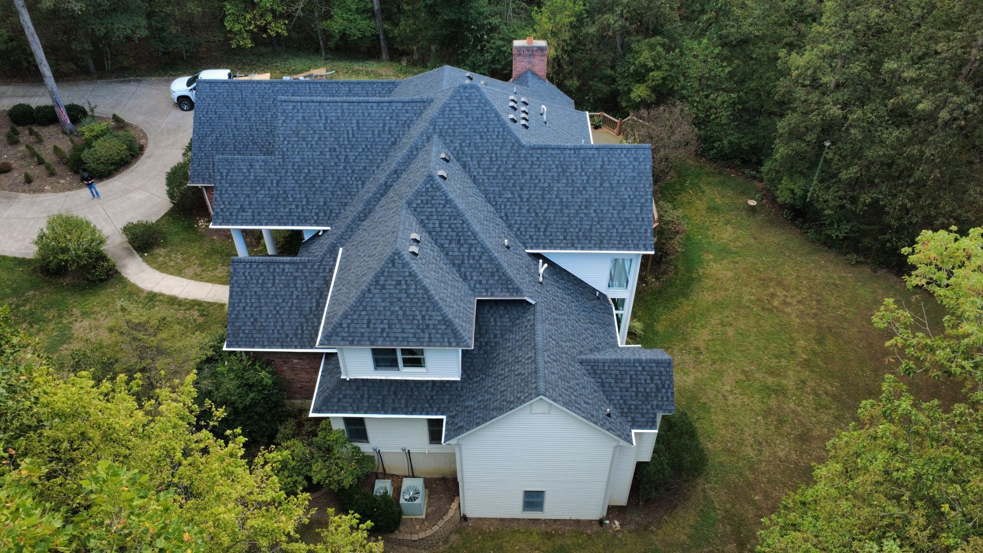 High-angle aerial view of a house with a dark shingled roof, set on a grassy lot surrounded by trees.