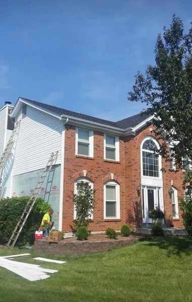 A worker in a high-visibility vest stands by a ladder against a two-story brick and white-sided house under a blue sky.