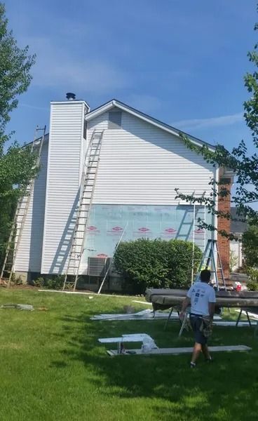 A worker walks toward a house with exposed siding underlayment and a ladder leaning against the chimney.
