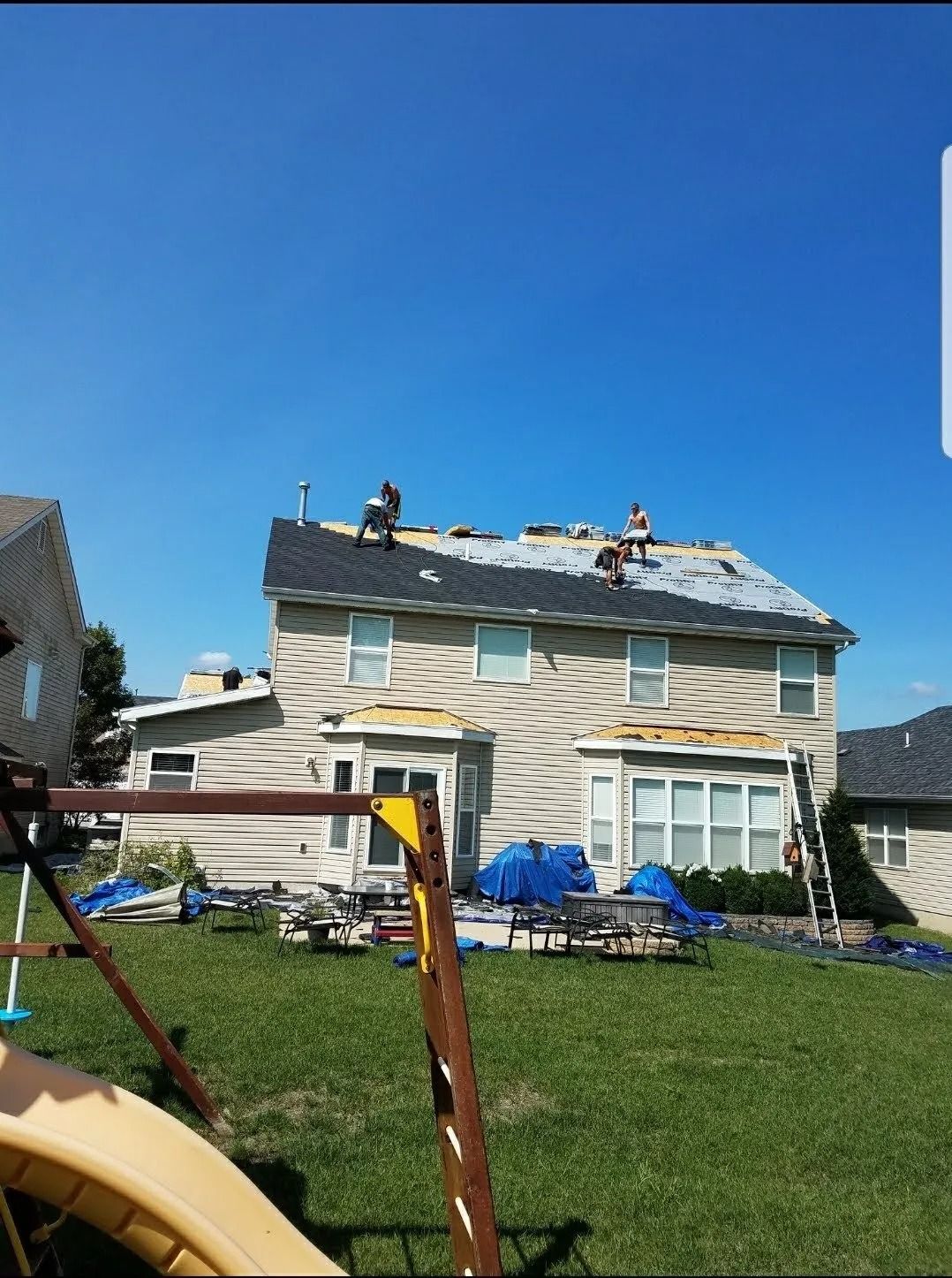 Workers install new shingles on the roof of a two-story beige house on a sunny day.