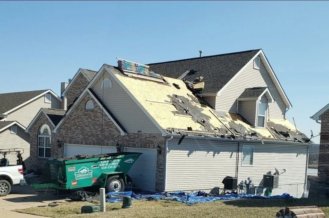 A two-story house with significant roof damage and exposed sheathing, with a green construction dumpster in the driveway.