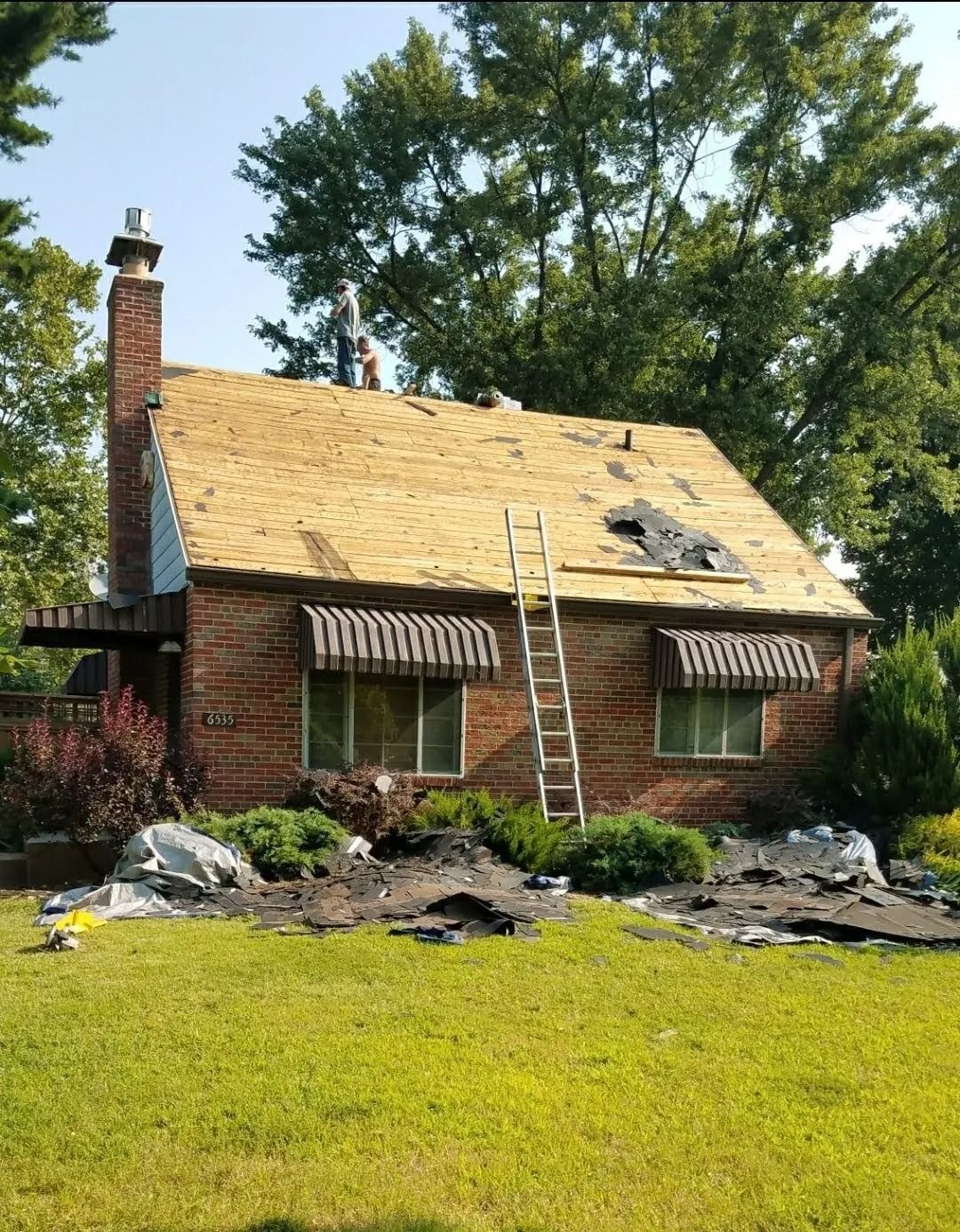 A brick house with a partially replaced shingle roof, a ladder leaning against it, and roof debris on the lawn.